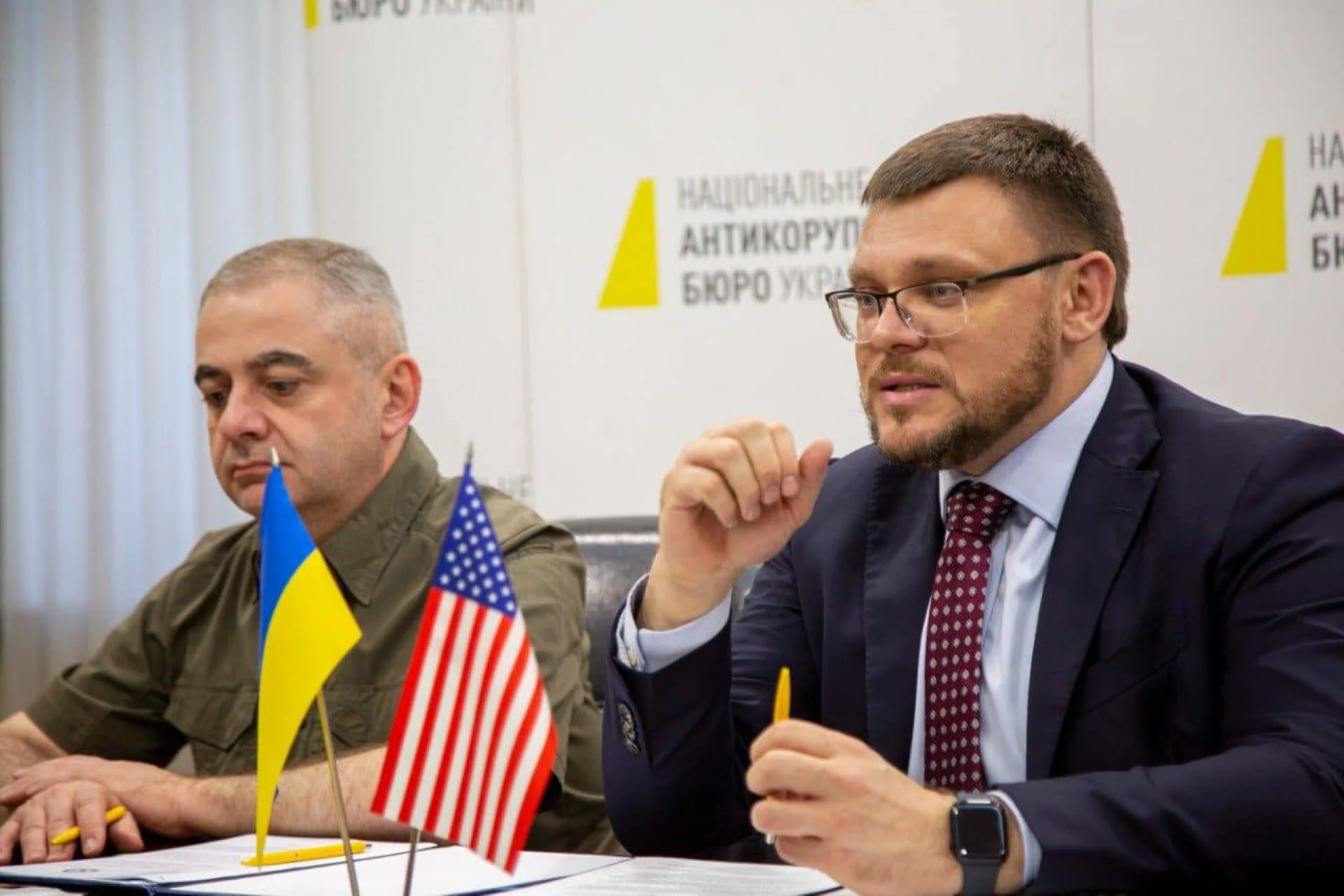 Кривонос у костюмі говорить на брифінгу НАБУ з прапорами України та США