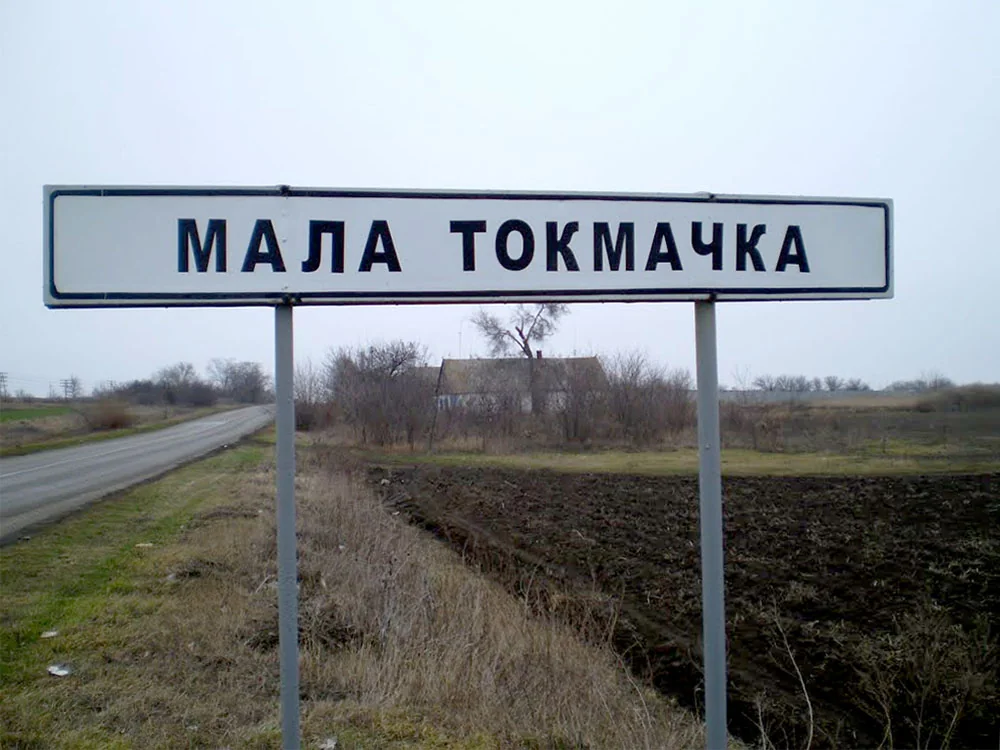 Дорожній знак «Мала Токмачка» Запорізької області на тлі села та полів.