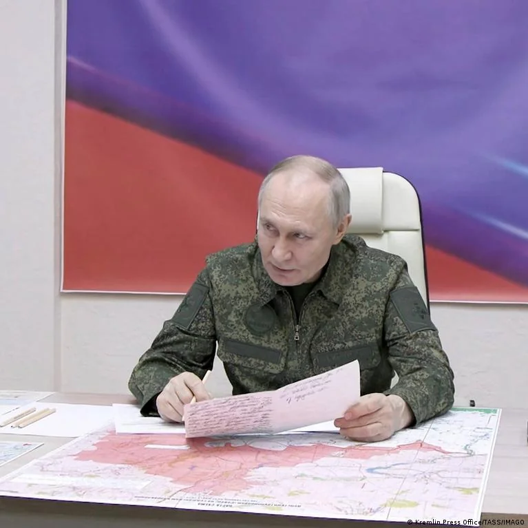 Володимир Путін у камуфляжі тримає документ над картою Росії на тлі прапора