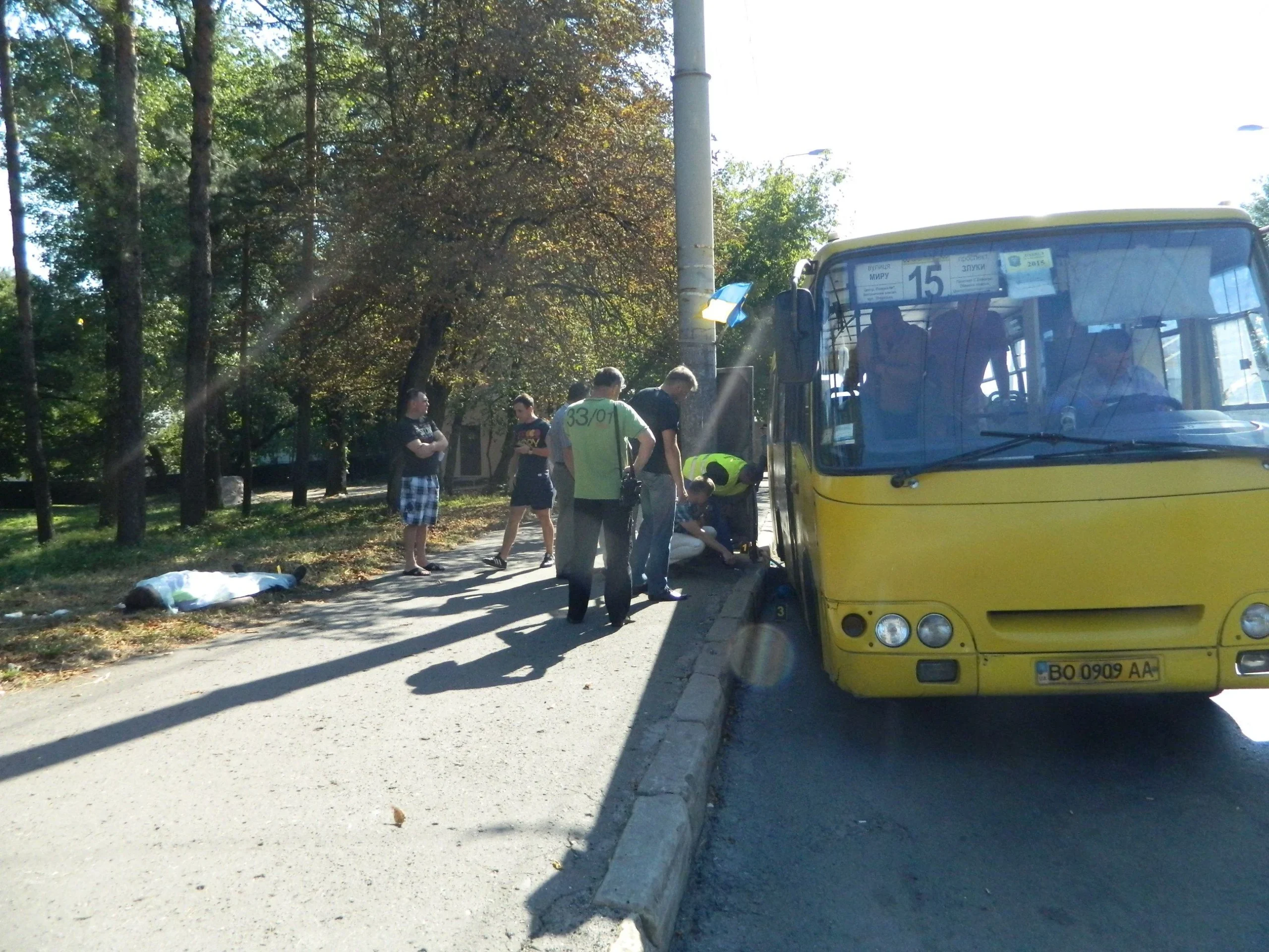 Жовтий автобус №15 біля тротуару, група людей і людина під білим покривалом
