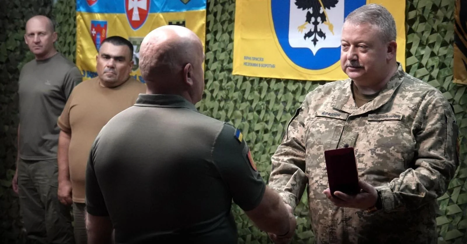 Військовий у камуфляжі вручає нагороду іншому солдату на фоні прапорів