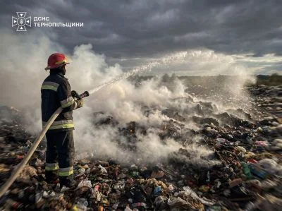 Пожежник у червоній касці гасить водою тліюче сміттєзвалище під хмарами