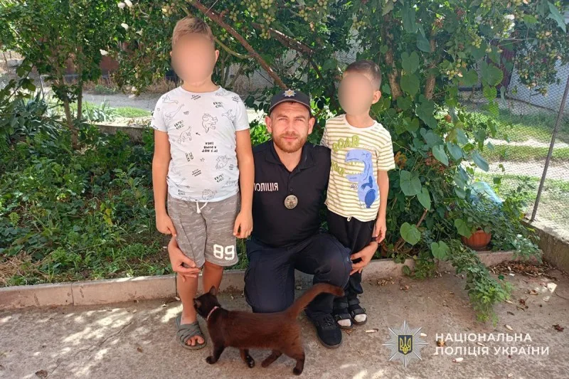Поліцейський навколішках з двома хлопчиками і коричневим котом біля кущів